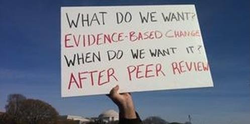 A protester holds a sign reading, "What do we want? Evidence-based change. When do we want it? After peer review," advocating for research-backed decision-making.