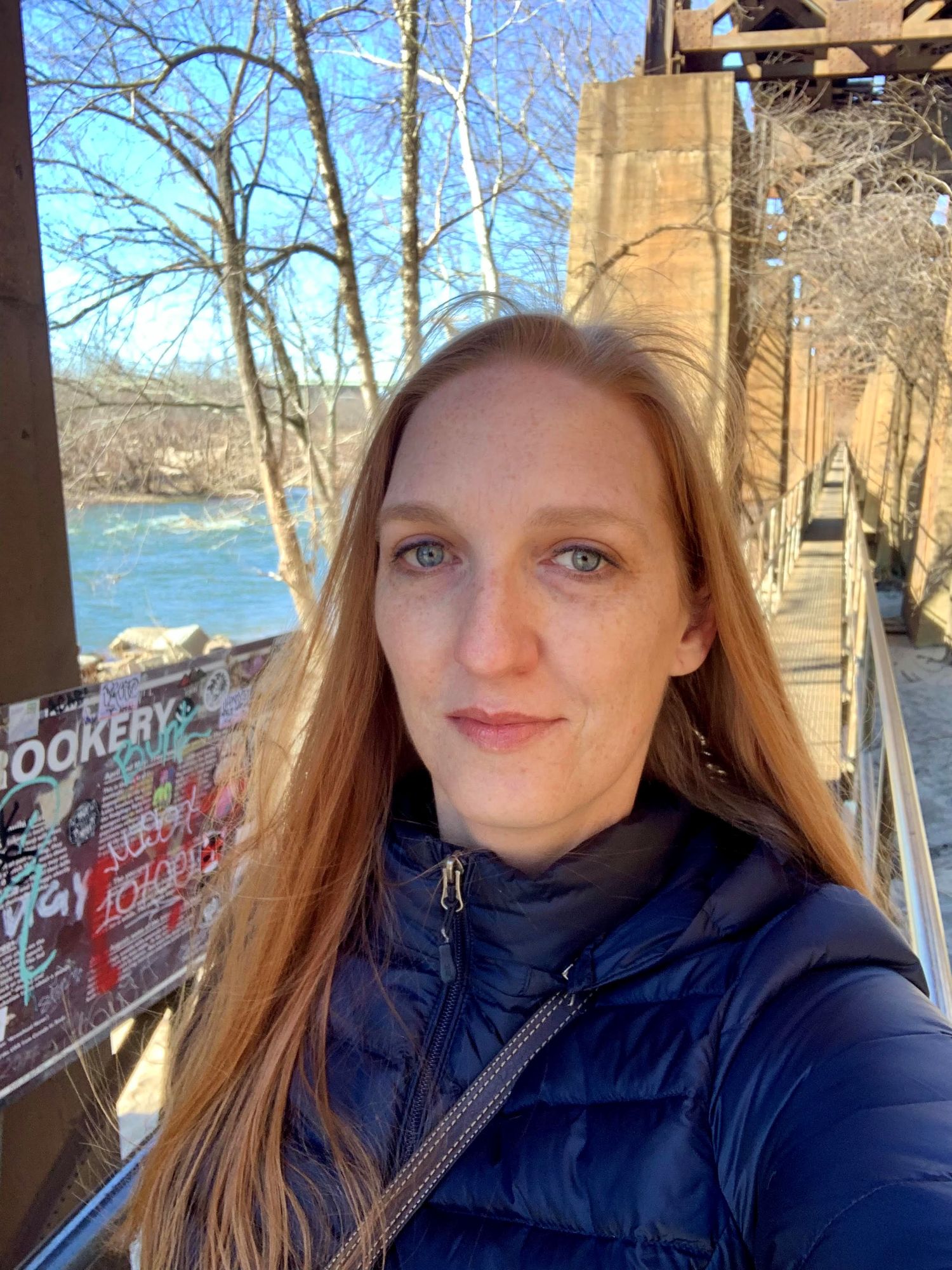 Jennifer Bronson stands on a walkway beside a river, framed by bare trees and industrial structures, evoking a tranquil outdoor scene.